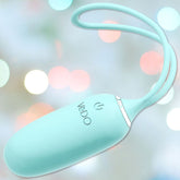 A turquoise wireless VeDO Kiwi Bullet Vibe, crafted in an egg shape with a flexible loop and power button, displays "VeDO" on its surface. This rechargeable bullet offers various vibration modes against a backdrop of soft, out-of-focus pastel lights.